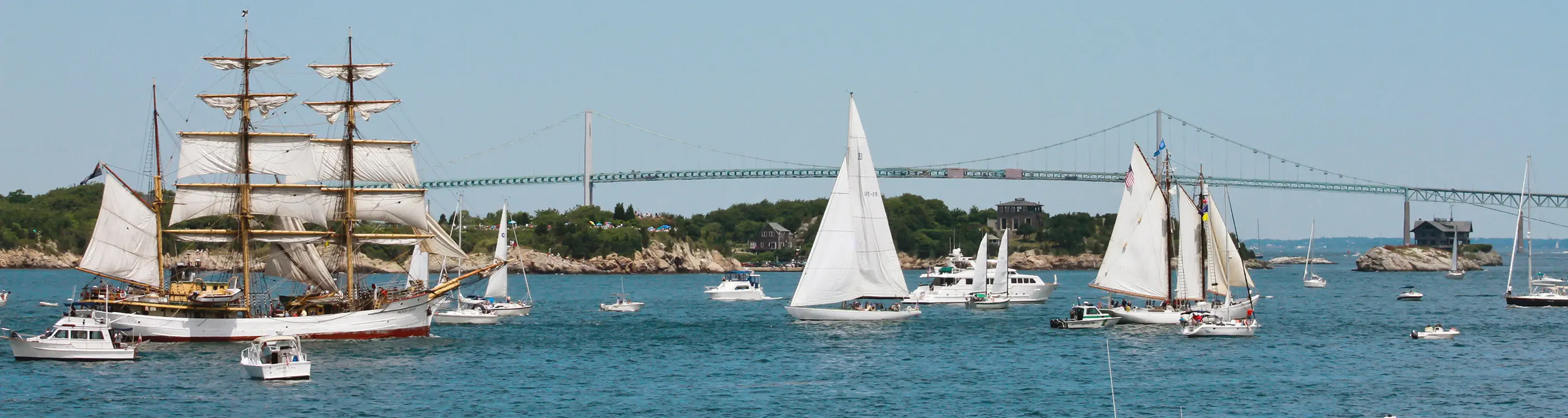 Tall ships and sailboats under sail on Narragansett Bay, passing beneath the Claiborne Pell Newport Bridge