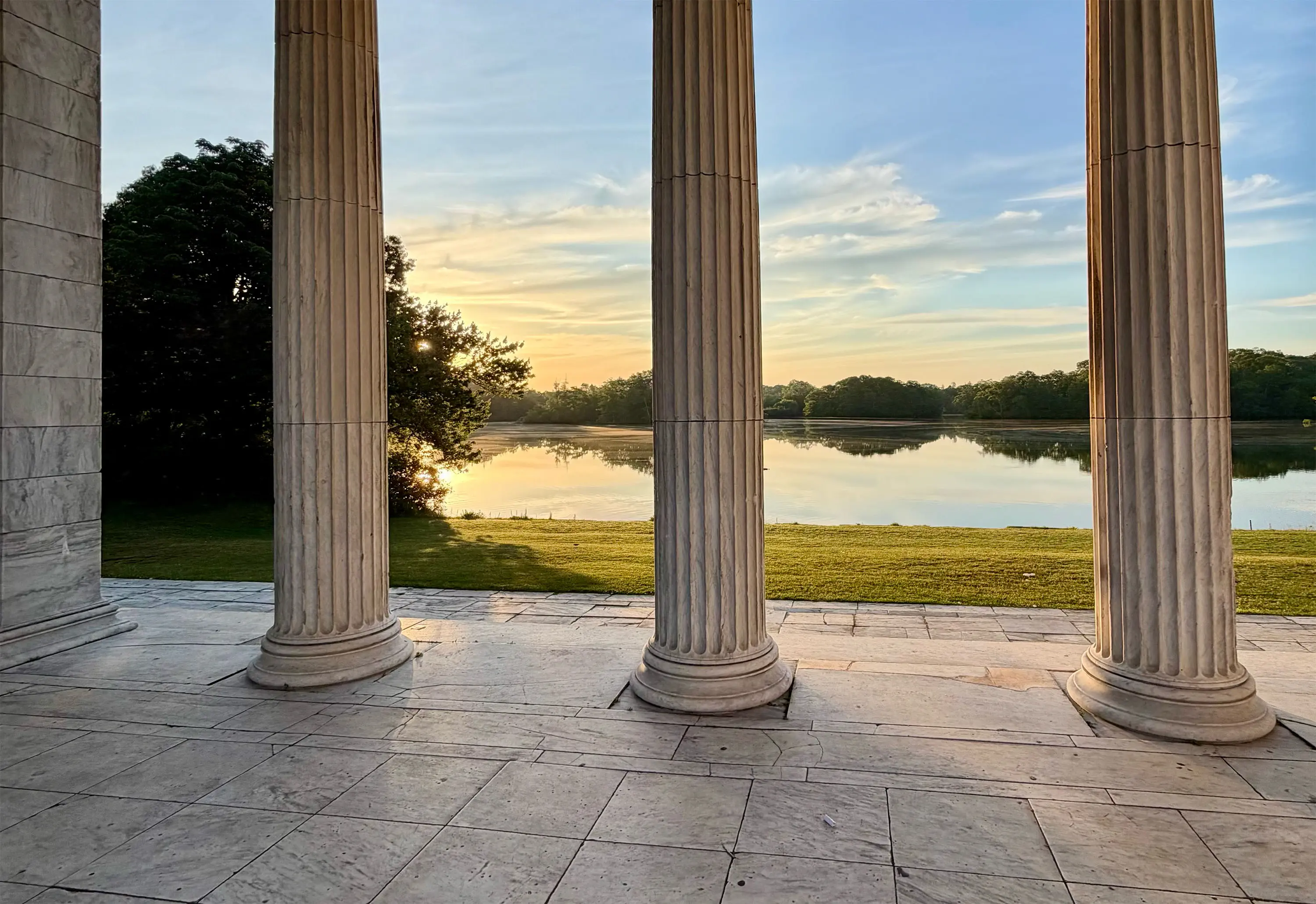 Sunrise seen between the marble columns of a colonnade, over a lawn and a still lake with trees on the far bank.