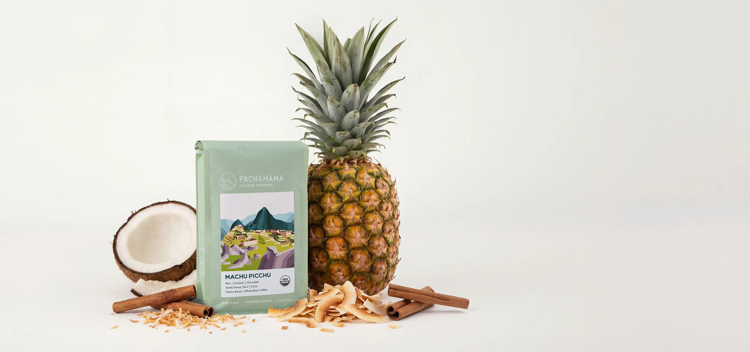 A bag of Machu Picchu Organic coffee beside a whole pineapple, a split coconut, toasted coconut flakes and cinnamon sticks